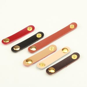 May include: Six rectangular leather strips in various colors including red, black, tan, beige, and dark brown. Each strip features two polished gold-tone metal snap buttons at the ends, providing a secure closure mechanism for these small leather items.