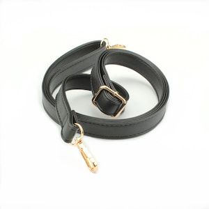 May include: A black adjustable bag strap made of smooth material with gold-toned metal hardware. The strap features a sliding buckle for length adjustment and two swivel lobster claw clasps at the ends for secure attachment to a bag or accessory.