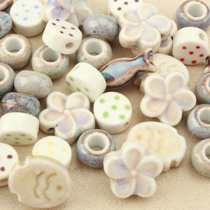 May include: A collection of ceramic beads in various shapes and finishes. The assortment includes flower-shaped beads with iridescent glazes, round beads with colourful polka dots, textured speckled beads, and fish-shaped beads in blue and tan tones.