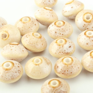 May include: A collection of round, disc-shaped ceramic beads with a glossy, cream-coloured glaze. Each bead features a central hole and a speckled, light brown pattern concentrated around the centre, creating a textured, spotted appearance.