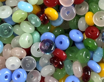 May include: A collection of colorful, translucent beads in various shapes and sizes. The beads are primarily donut-shaped, with a few oval and irregular forms. Colors include red, yellow, green, blue, and clear. These beads are suitable for jewelry making and crafts.