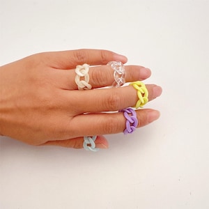 Peut inclure: Une main présente cinq bagues en forme de chaîne de différentes couleurs. Les bagues sont beige clair, transparentes, jaunes, violettes et bleu clair. Les bagues sont en matériau solide et ont un design moderne et épais. Les bagues sont un accessoire de mode.