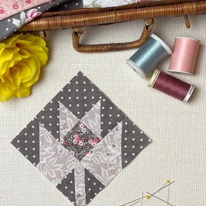 Puede incluir: Un bloque de colcha de tela gris y blanco con un patr&oacute;n floral en el centro. El bloque est&aacute; rodeado de tela gris con lunares blancos. Hay alfileres de costura y carretes de hilo sobre una superficie blanca.