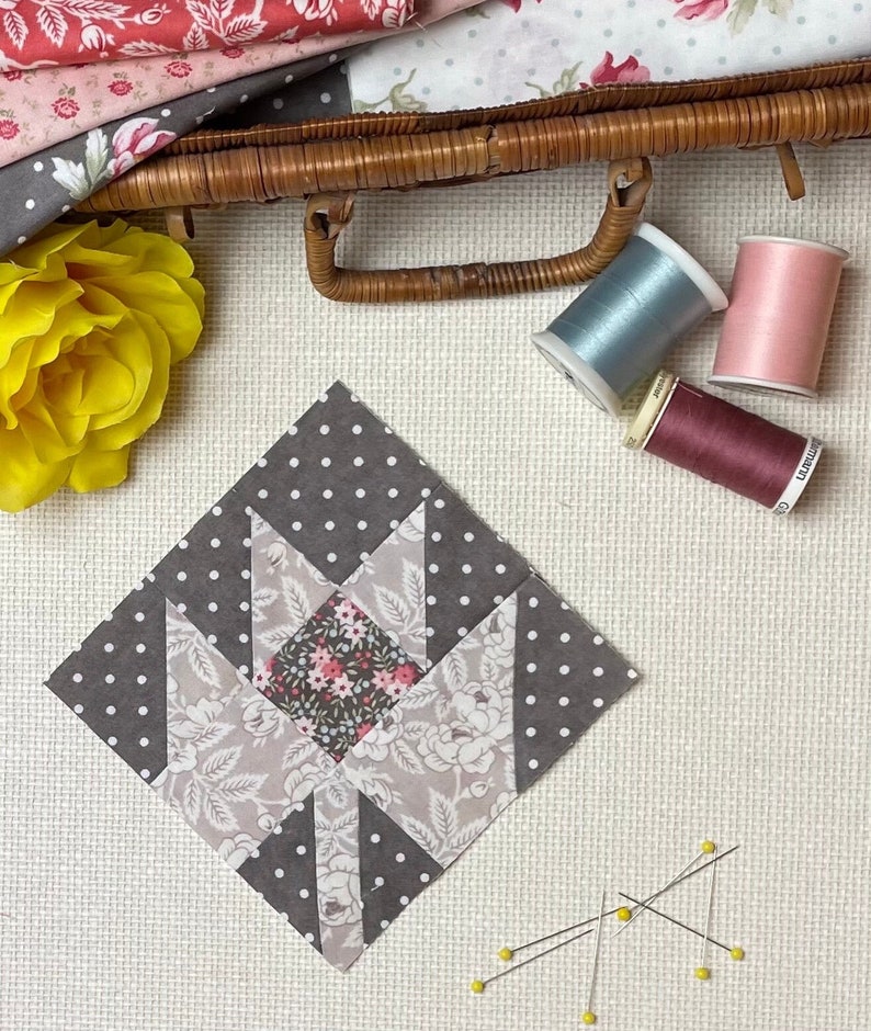 Puede incluir: Un bloque de colcha de tela gris y blanco con un patr&oacute;n floral en el centro. El bloque est&aacute; rodeado de tela gris con lunares blancos. Hay alfileres de costura y carretes de hilo sobre una superficie blanca.