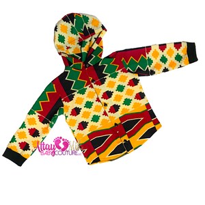May include: A button-down hooded shirt with a vibrant geometric pattern in red, yellow, green, and black. The shirt features long sleeves with black cuffs and a matching hood. The text "Attay Mik Baby Couture LLC" is printed on the shirt.