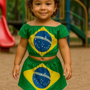 以下が含まれることがあります： ブラジル国旗をあしらったグリーンのツーピースの衣装を着た幼い子供。トップスはオフショルダーのクロップトップで、スカートはミニスカートです。衣装には、青い円と星が付いた黄色のひし形と「ORDEM E PROGRESSO」の文字が描かれています。