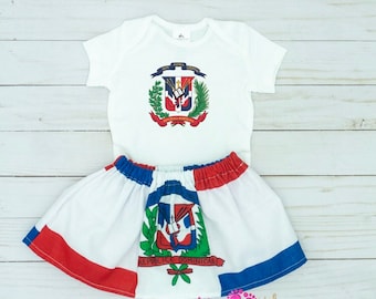 May include: A white baby outfit set featuring the Dominican Republic coat of arms on the short-sleeved top and skirt. The skirt has red and blue trim. The outfit is displayed on a white wooden surface.