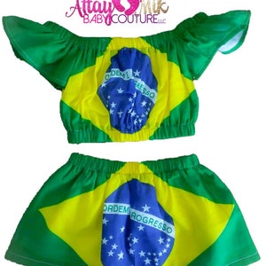 以下が含まれることがあります： ブラジル国旗のデザインが施された2ピースのベビー服。セットには、フリル袖のオフショルダーのトップスと、お揃いのスカートが含まれています。トップスとスカートは、青いエンブレムと緑の縁取りのある黄色のセンターを備えています。