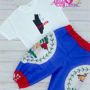 Può includere: Una maglietta bianca con una grafica nera e rossa di una mappa di un paese e il nome "Travis". L'outfit include pantaloni blu con un bordo rosso e un emblema del paese. L'outfit è progettato per bambini.