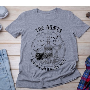 May include: A heather gray short-sleeve t-shirt featuring a black graphic print. The design includes a central bottle, a cauldron, a cocktail glass, and bats, with the text: THE AUNTS MIDNIGHT MARGARITAS since 1998 Someone left it on the porch.