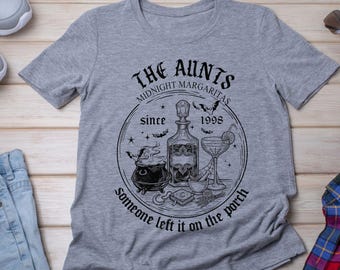 May include: A heather gray short-sleeve t-shirt featuring a black graphic print. The design includes a central bottle, a cauldron, a cocktail glass, and bats, with the text: THE AUNTS MIDNIGHT MARGARITAS since 1998 Someone left it on the porch.