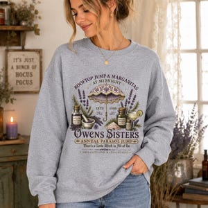 May include: A heather gray crewneck sweatshirt featuring a graphic print with a purple parasol, lavender sprigs, apothecary bottles, and a margarita. The text reads: ROOFTOP JUMP & MARGARITAS AT MIDNIGHT, OWENS SISTERS, ANNUAL PARASOL JUMP.