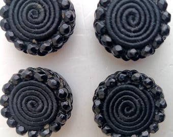 Può includere: Quattro bottoni neri con un motivo a spirale al centro, circondati da un bordo di perline. I bottoni sono rotondi e sembrano essere fatti di tessuto e perline. I bottoni sono su una superficie bianca.