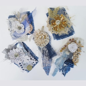 Puede incluir: Cinco broches de tela hechos a mano con estética vintage. Cada broche presenta una base de mezclilla azul o arpillera, adornada con encaje, lentejuelas, cuentas y flores de tela en tonos blanco, crema y dorado. Los broches varían en tamaño y diseño.