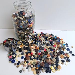 Puede incluir: Un frasco grande de vidrio transparente lleno de un vasto surtido de botones de varios colores, formas y tamaños, acompañado de un pequeño recipiente de metal y un gran montón de botones sueltos esparcidos por la superficie frente al frasco.