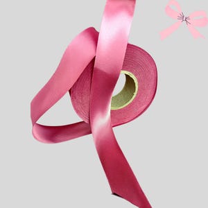 May include: A roll of pink satin ribbon with a glossy finish. The ribbon is partially unrolled, revealing the spool's cardboard core. A small illustration of a pink bow is in the upper right corner. Suitable for crafts, gift wrapping, and decorative use.