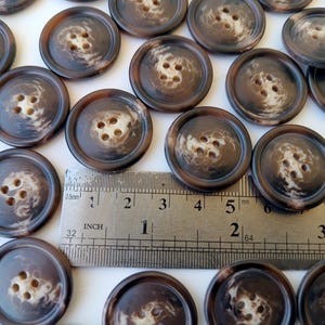 Könnte beinhalten: Eine Sammlung von 20 braunen und weißen, marmorierten Kunststoffknöpfen mit jeweils vier Löchern. Die Knöpfe sind in einem zufälligen Muster auf einer weißen Oberfläche angeordnet. Ein Lineal ist unter den Knöpfen platziert und zeigt Maße in Zoll und Millimetern.