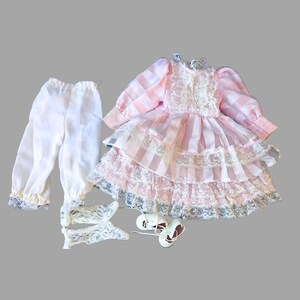 Op de afbeelding: Een poppenoutfit bestaande uit een roze en wit geruite jurk met lange mouwen en witte kanten afwerking, een witte broek met elastische taille en kanten boorden, een paar witte kanten sokken en een paar witte schoenen met dunne bandjes.