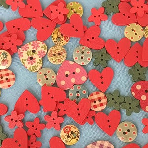 Puede incluir: Una colección de botones de madera surtidos en varias formas, incluyendo corazones, flores y círculos. Los botones presentan diseños rojos, verdes, amarillos y estampados con dos o cuatro agujeros para coser, esparcidos sobre una superficie de tela azul claro.
