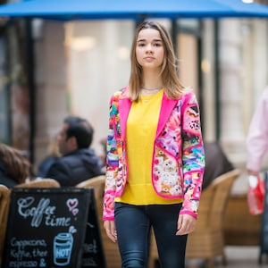 Può includere: Una persona giovane indossa un blazer rosa con una stampa colorata di animali dei cartoni animati e dolci. Il blazer è indossato sopra un maglione giallo e jeans blu.