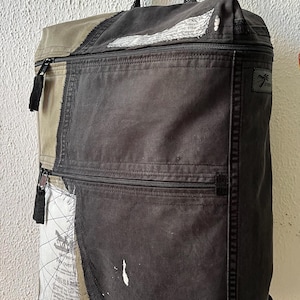 May include: A dark gray backpack with multiple zippered compartments and a top handle. The backpack features a patchwork design with sections of tan and white fabric with printed text. Black tassels hang from the zippers.