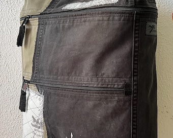 May include: A dark gray backpack with multiple zippered compartments and a top handle. The backpack features a patchwork design with sections of tan and white fabric with printed text. Black tassels hang from the zippers.