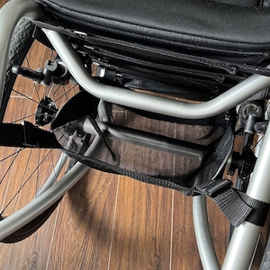 Peut inclure: Un fauteuil roulant argent&eacute; avec un sac de rangement en maille noire fix&eacute; au cadre. Le fauteuil roulant a un si&egrave;ge noir et un repose-pieds noir.