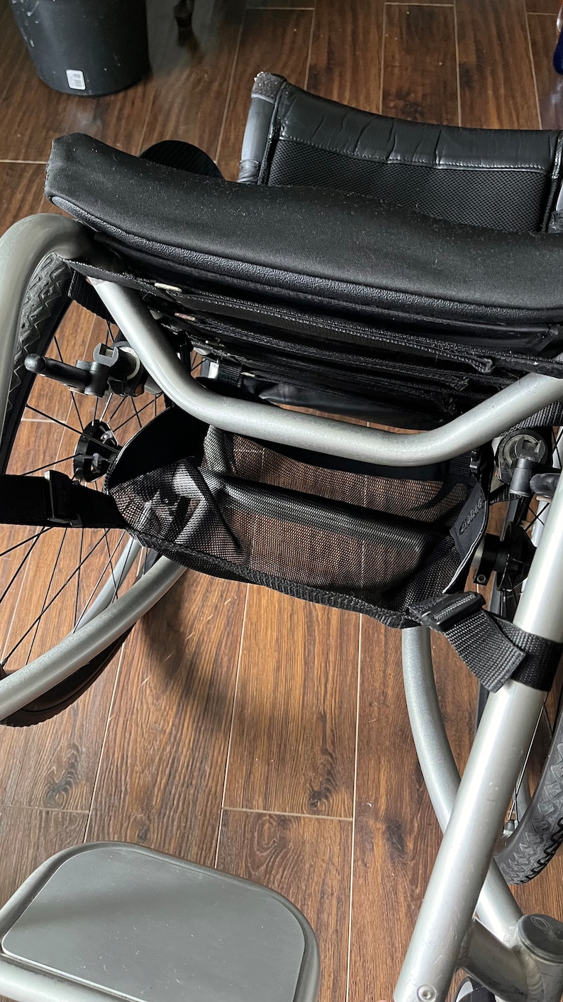 Peut inclure: Un fauteuil roulant argent&eacute; avec un sac de rangement en maille noire fix&eacute; au cadre. Le fauteuil roulant a un si&egrave;ge noir et un repose-pieds noir.