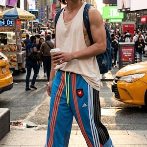 Op de afbeelding: Een persoon draagt een crèmekleurige tanktop, een blauwe trainingsbroek met oranje en witte strepen en witte sneakers. De broek heeft een rode patch met de letters "vi". Een denim rugzak zit op de rug van de persoon.