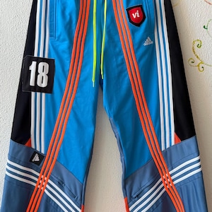 May include: Blue and black track pants with orange stripes and white accents. The pants feature the number "18" on a black patch and a red "vi" logo. The Adidas logo is visible.