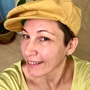 May include: A woman with short brown hair is wearing a yellow newsboy cap. She is smiling and looking at the camera. She is wearing a light green shirt.