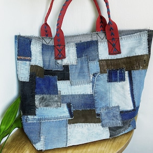 May include: A large tote bag made from patchwork denim fabric. The bag has red braided handles and is made up of various shades of blue denim.