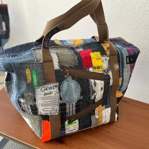 May include: A brown and blue patchwork tote bag made from recycled denim. The bag has a zipper closure and features various patches and labels, including "Gears denim" and "F.I.D.A."