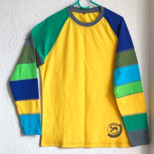 Peut inclure: T-shirt à manches longues jaune avec une manche rayée verte, bleue et jaune. Le t-shirt a un col rond gris et un logo qui dit "Dragons Amsterdam Recycled Skateboard Collection".