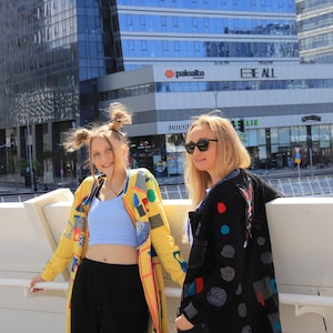 Puede incluir: Dos personas con chaquetas coloridas se paran frente a un edificio. La persona de la izquierda lleva una chaqueta amarilla con parches y un top corto azul. La persona de la derecha lleva una chaqueta negra con parches coloridos y jeans azules. El edificio tiene un letrero que dice "paloalto" y "BE ALL".