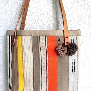 May include: A striped tote bag with tan, yellow, orange, and white vertical stripes. The bag has brown leather handles and two small pom-poms attached. The text "BERRY FRAGRANCES BURBER" is printed on the bag.