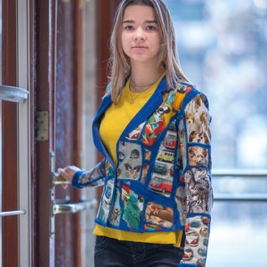 Puede incluir: Una persona joven lleva una chaqueta azul con un diseño de patchwork colorido que presenta animales, insectos y vehículos. La chaqueta tiene una camisa amarilla debajo y pantalones vaqueros azules.
