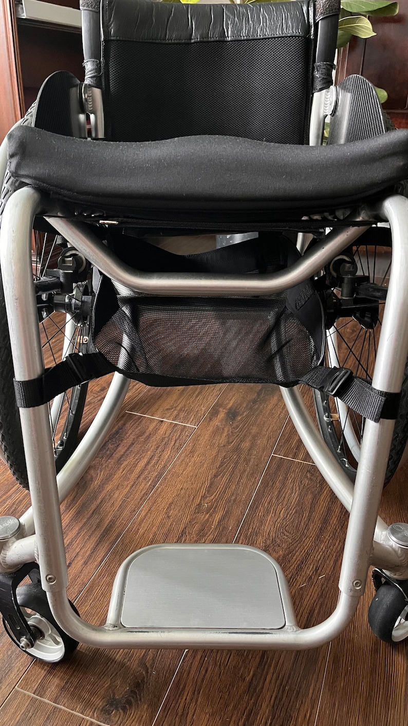 Peut inclure: Un fauteuil roulant argent&eacute; avec un si&egrave;ge et un dossier en tissu noir, un sac de rangement en maille noire et un repose-pieds argent&eacute;.