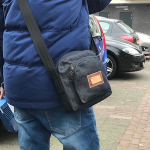 May include: A person wearing a blue puffer jacket and blue jeans is carrying a black denim crossbody bag with a brown leather patch. The bag has a zipper closure and a strap.