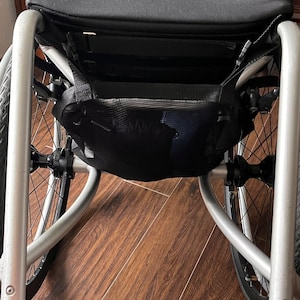 Peut inclure: Un fauteuil roulant argent&eacute; avec un sac de rangement en maille noire fix&eacute; &agrave; l'arri&egrave;re. Le fauteuil roulant a un repose-pieds et un si&egrave;ge rembourr&eacute; noir.