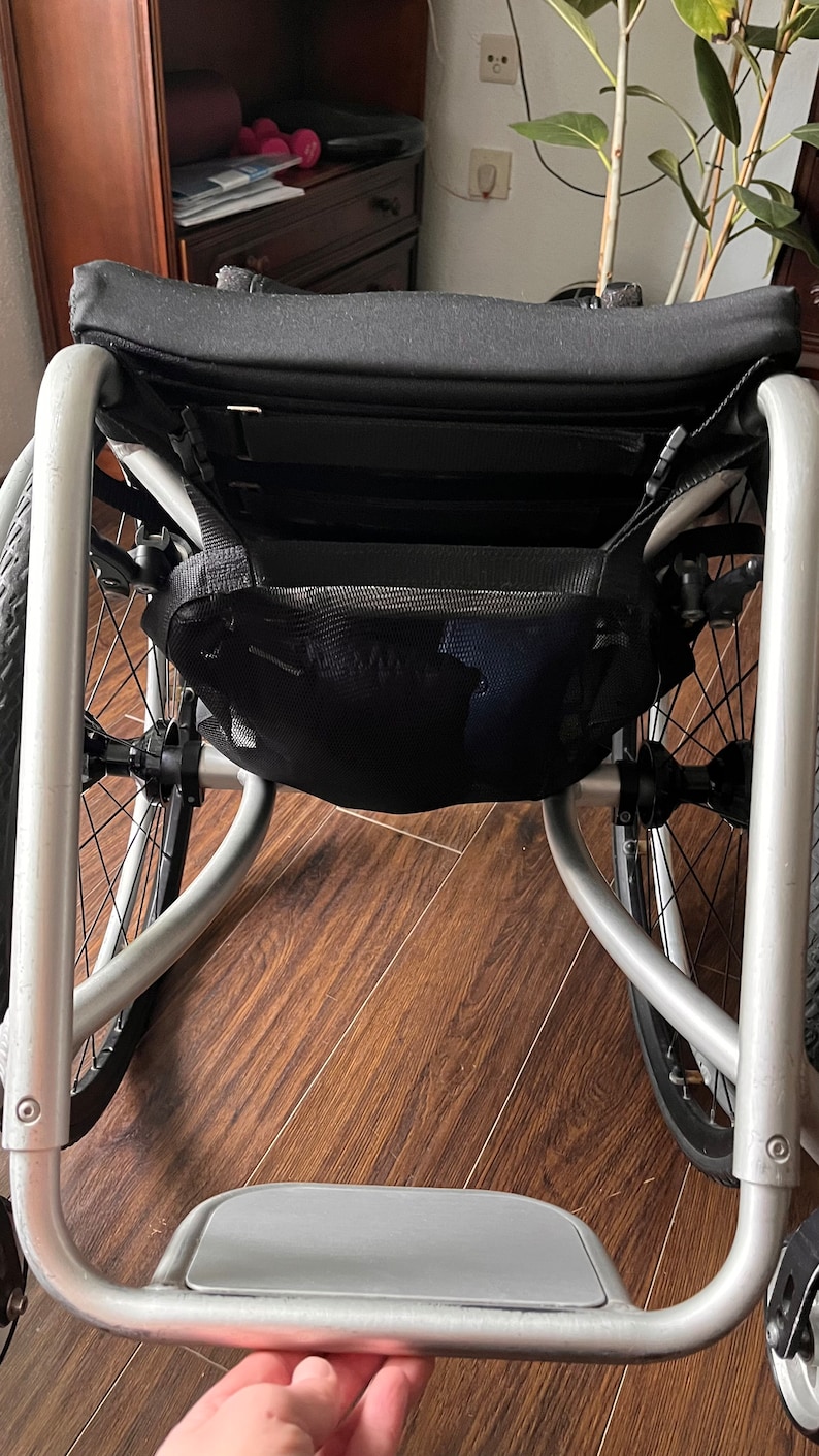 Peut inclure: Un fauteuil roulant argent&eacute; avec un sac de rangement en maille noire fix&eacute; &agrave; l'arri&egrave;re. Le fauteuil roulant a un repose-pieds et un si&egrave;ge rembourr&eacute; noir.