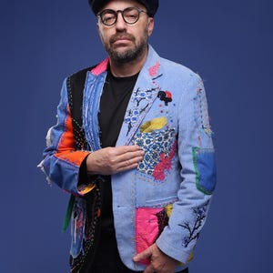 May include: A man wearing a blue, multi-coloured patchwork jacket with a black shirt and a dark blue newsboy cap. He is wearing glasses and has a beard.