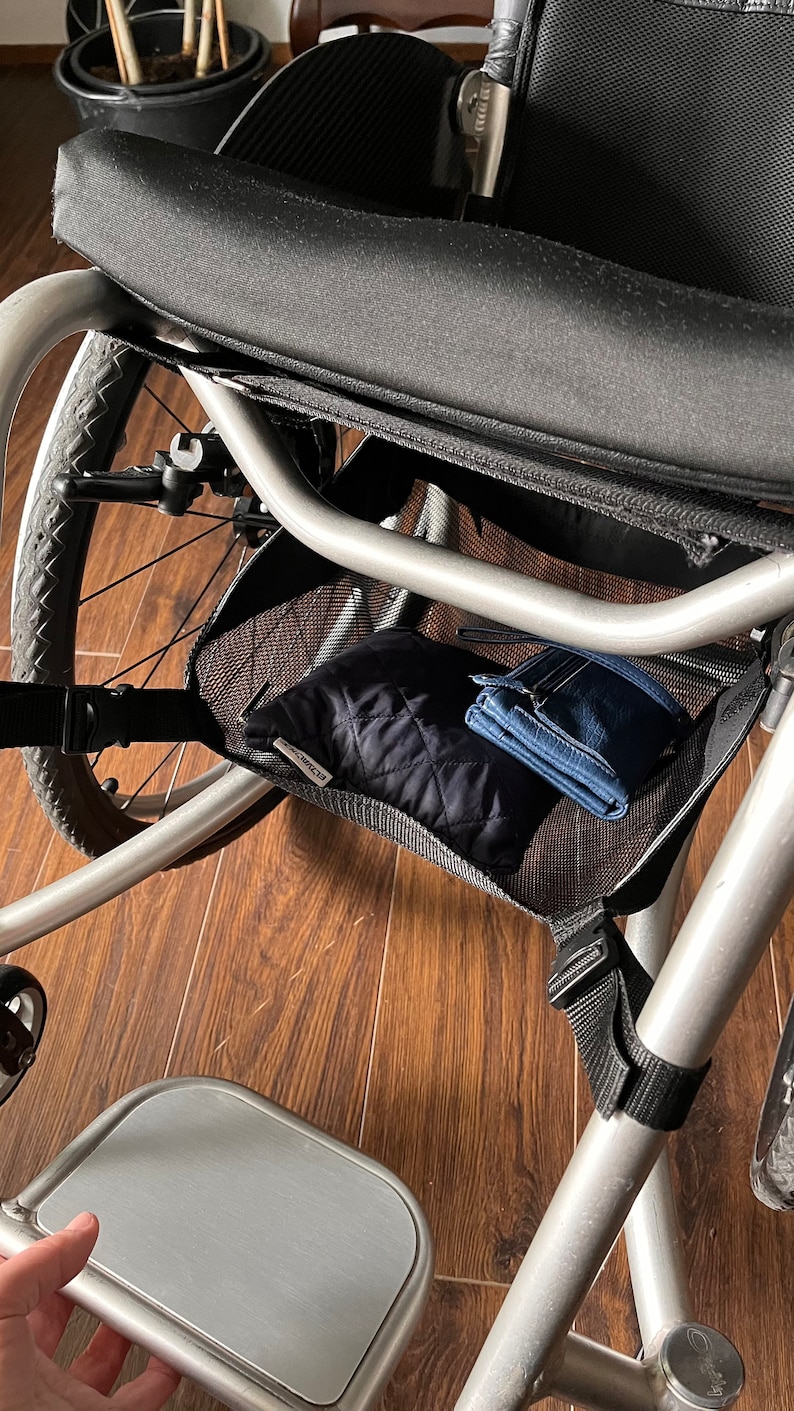 Peut inclure: Un fauteuil roulant argent&eacute; avec un sac de rangement en maille noire sous le si&egrave;ge. Le sac contient une pochette matelass&eacute;e noire et un portefeuille bleu. Le fauteuil roulant a un repose-pieds argent&eacute;.