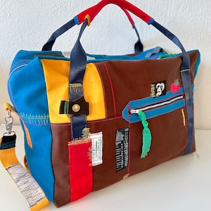 May include: A multi-coloured duffel bag made from recycled materials. The bag has a brown, blue, yellow, and red patchwork design. The bag has a zipped pocket and a top handle.