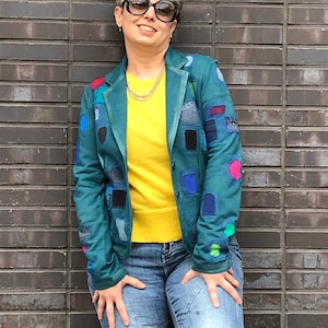 Pode incluir: Blazer azul-petróleo com detalhes de patchwork coloridos, usado sobre um suéter amarelo e jeans azuis. O blazer tem um corte clássico com fecho de botões. O patchwork inclui várias formas e cores, adicionando um toque artístico único.