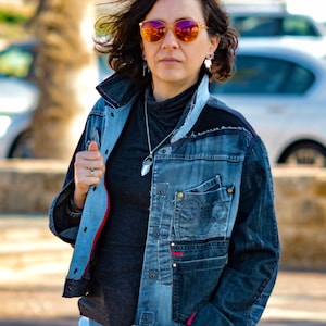 Puede incluir: Una mujer con una chaqueta vaquera azul con ribete rojo y una camiseta de cuello alto negra. Lleva gafas de sol naranjas y un collar de plata con un colgante de cristal.