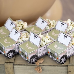 May include: A collection of small, camouflage-patterned jeep-shaped boxes filled with popcorn. Each box features a white label and a wing design. The boxes are displayed on a wooden crate, suitable for a themed party.