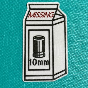 May include: A white sticker in the shape of a milk carton with the word "MISSING" in red at the top. The carton features a black graphic of a bullet and the text "10mm" in black.
