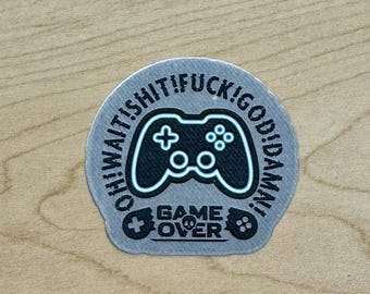 May include: A round sticker with a black and white design. The sticker features a game controller in the center, surrounded by the phrases "OH! WAIT! SHIT! FUCK! GOD! DAMN!". Below the controller, the text reads "GAME OVER".