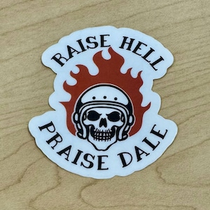 May include: A white sticker with a black skull wearing a helmet, surrounded by orange flames. The sticker has the words "RAISE HELL" and "PRAISE DALE" in black, curved around the top and bottom of the design.